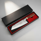 A Japanese-style kitchen knife with a patterned steel blade and a dark wood handle, presented in an open black gift box with a red interior. The box is set on a dark textured surface with a warm spotlight, and three raw beef cubes and rosemary sprigs are placed nearby.