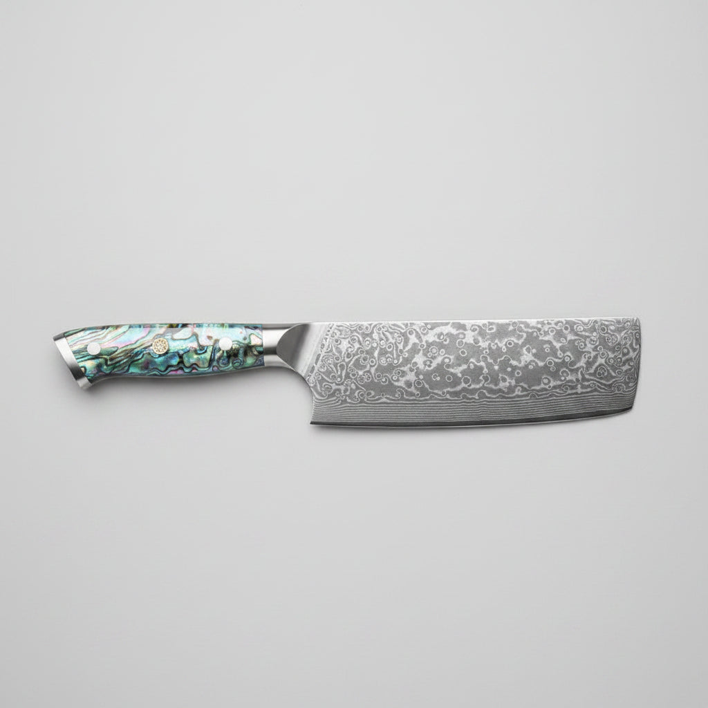 A Nakiri knife with a patterned Damascus steel blade and an iridescent abalone shell handle, displayed diagonally on a wooden cutting board with sliced cucumbers, kale, and a small bowl of radishes.
