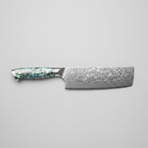 A Nakiri knife with a patterned Damascus steel blade and an iridescent abalone shell handle, displayed diagonally on a wooden cutting board with sliced cucumbers, kale, and a small bowl of radishes.