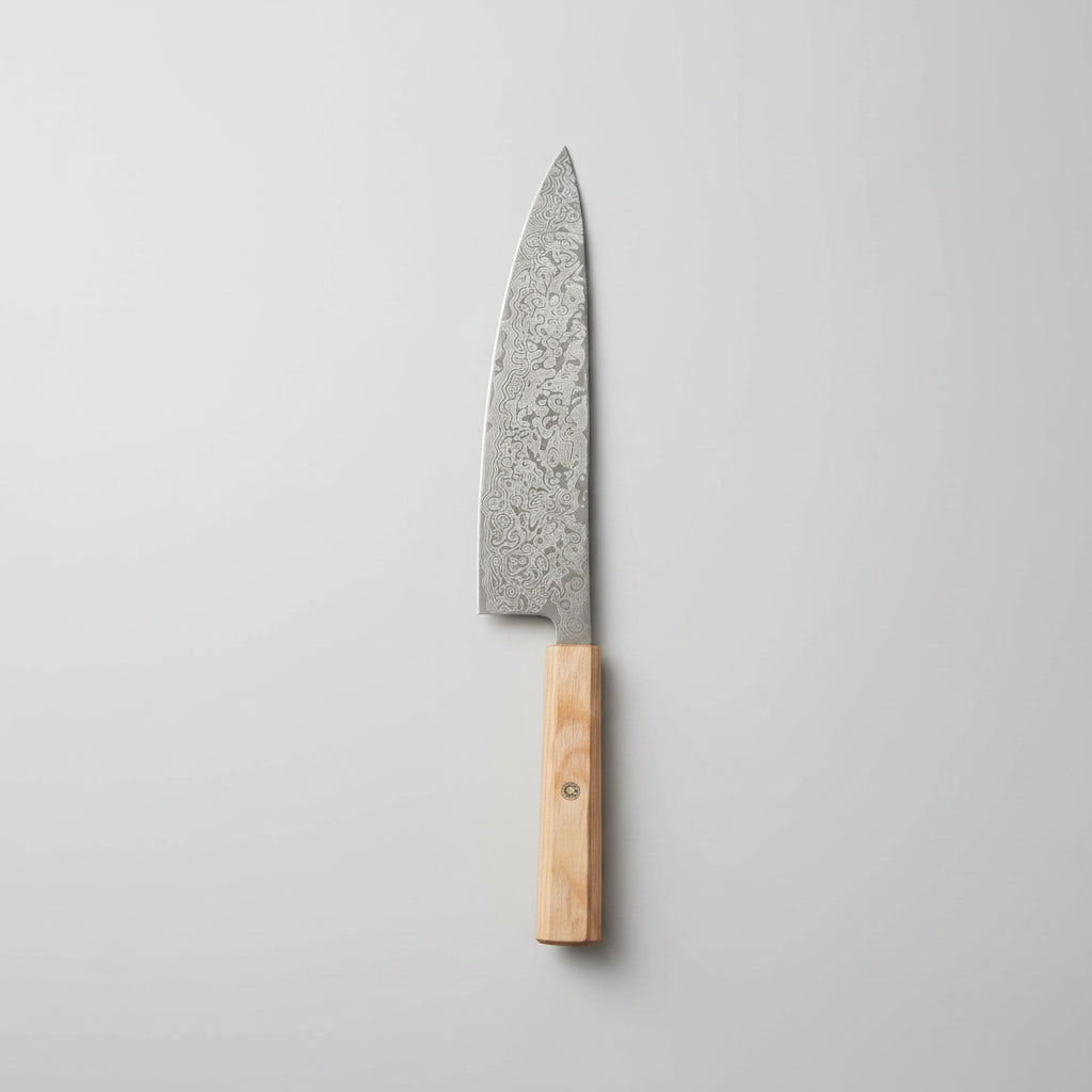 A chef's knife with a patterned steel blade and a light wood handle rests diagonally on a wooden cutting board, surrounded by fresh rosemary, garlic cloves, peppercorns, coarse salt, and a cloth on a rustic wooden table.