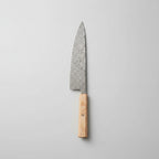 A chef's knife with a patterned steel blade and a light wood handle rests diagonally on a wooden cutting board, surrounded by fresh rosemary, garlic cloves, peppercorns, coarse salt, and a cloth on a rustic wooden table.