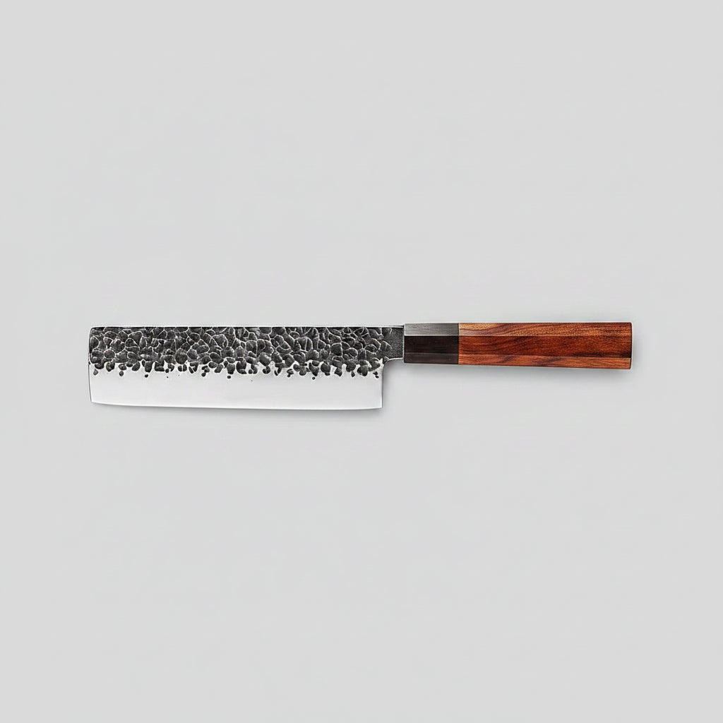 A kitchen knife with a hammered steel blade and a reddish-brown wooden handle, placed diagonally on a dark wooden cutting board surrounded by sliced red chilies, green bok choy, garlic, and a cloth.