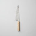 A kitchen knife with a patterned steel blade and a light wood handle resting on a wooden cutting board, surrounded by fresh rosemary, thyme, garlic, lemon slices, and a beige cloth on a marble surface.