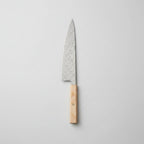 A kitchen knife with a patterned steel blade and a light wood handle resting on a wooden cutting board, surrounded by fresh rosemary, thyme, garlic, lemon slices, and a beige cloth on a marble surface.