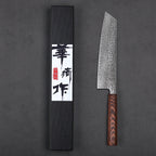 A Japanese-style chef's knife with a hammered Damascus steel blade and a dark brown wood handle, displayed next to its black presentation box with a white label featuring black calligraphy.