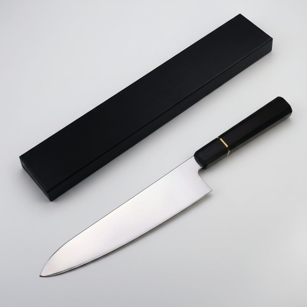 A professional chef's knife with a polished silver stainless steel blade and a black handle accented by a thin gold band, shown next to its sleek black presentation box on a white background.