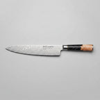 A chef's knife with a Damascus steel blade and a black handle with a peach-colored accent, displayed on a dark wooden table next to a raw steak on a wooden cutting board, with onions, avocado, and peppercorns.