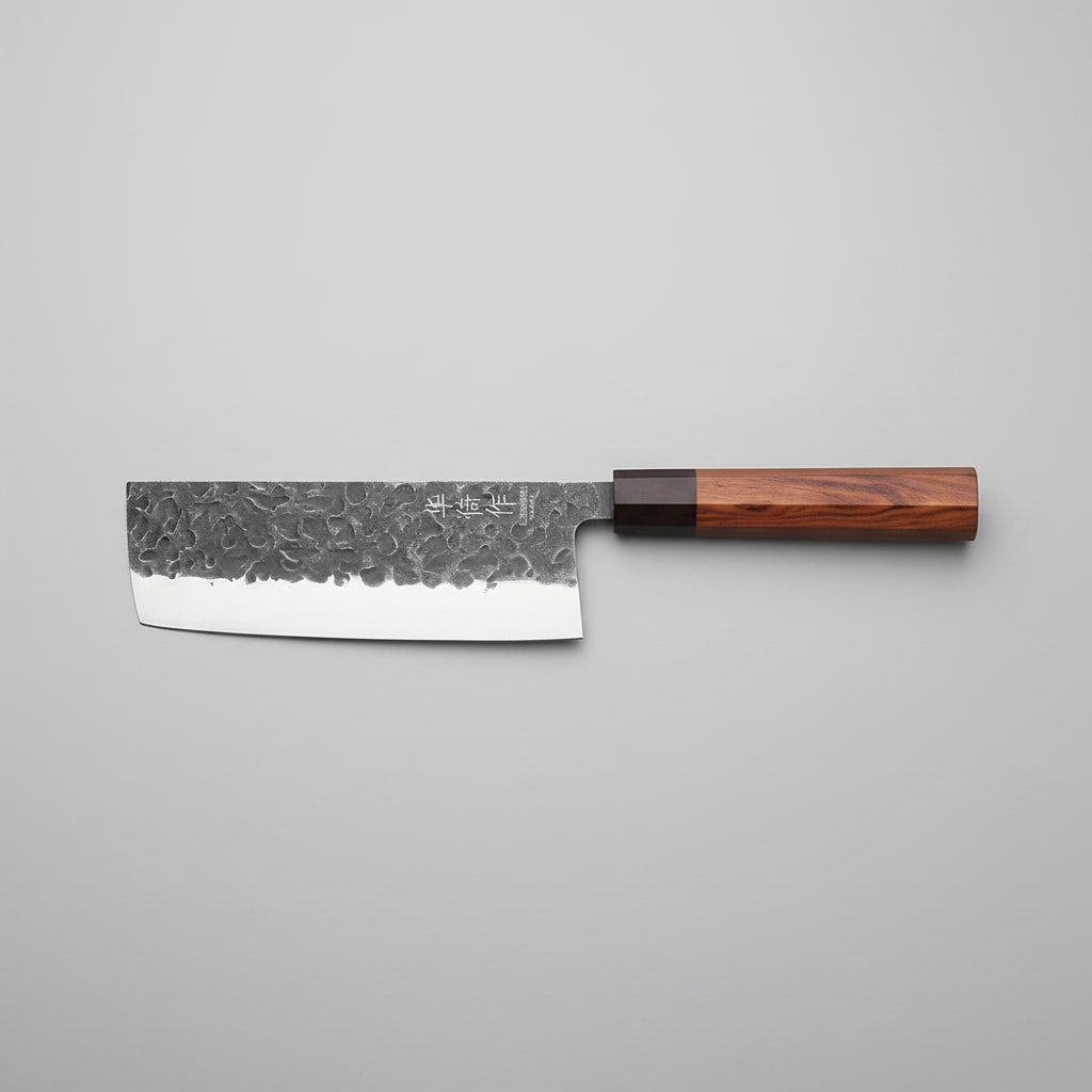 A Japanese-style kitchen knife with a hammered steel blade and a reddish-brown wooden handle, resting on a wooden cutting board surrounded by fresh purple cabbage, basil leaves, lemon slices, and a small red onion on a dark rustic table.
