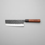 A Japanese-style kitchen knife with a hammered steel blade and a reddish-brown wooden handle, resting on a wooden cutting board surrounded by fresh purple cabbage, basil leaves, lemon slices, and a small red onion on a dark rustic table.