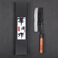 A Japanese-style kitchen knife with a hammered black blade and a brown wooden handle, displayed next to its black presentation box with white and red calligraphy on a dark gray background.