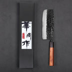 A Japanese-style kitchen knife with a hammered black blade and a brown wooden handle, displayed next to its black presentation box with white and red calligraphy on a dark gray background.