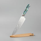 A Damascus steel chef knife with a patterned silver blade and a blue-green marbled handle, shown next to its light brown wooden sheath on a wooden cutting board. Text overlays on the left read '67 Layers Damascus Steel', 'Razor Sharp', and 'Chef Knife'.